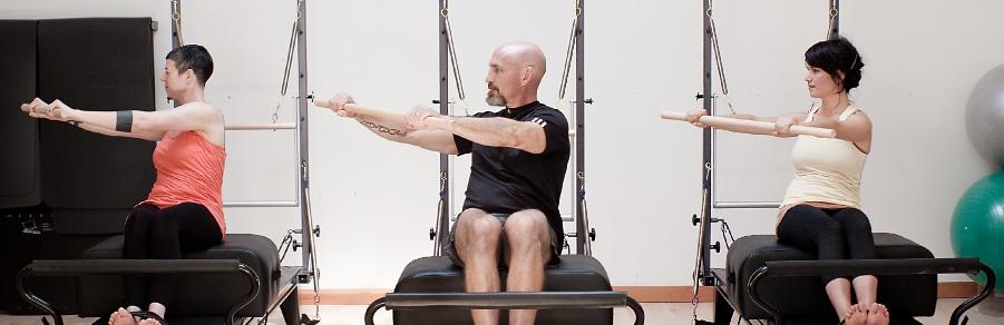 Three clients seated on Pilates chairs, arms extended forward on tower bars