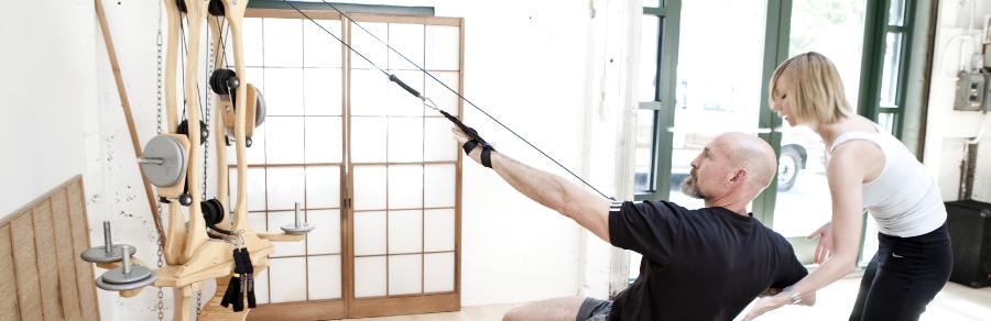 Trainer assisting a client reaching along the cables of a wooden GYROTONIC Pulley Tower